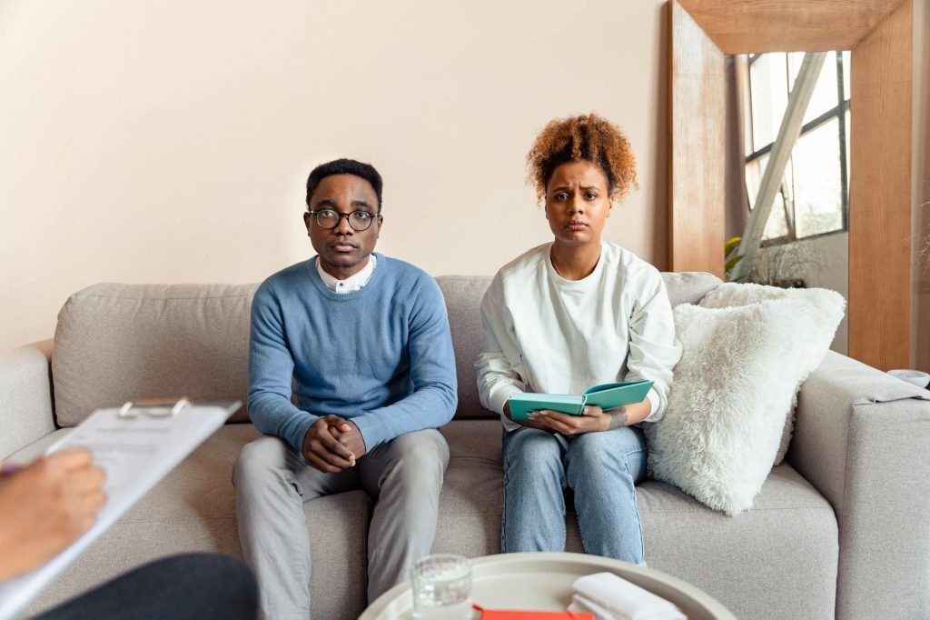 A couple sits on a sofa during a therapy session, appearing concerned.